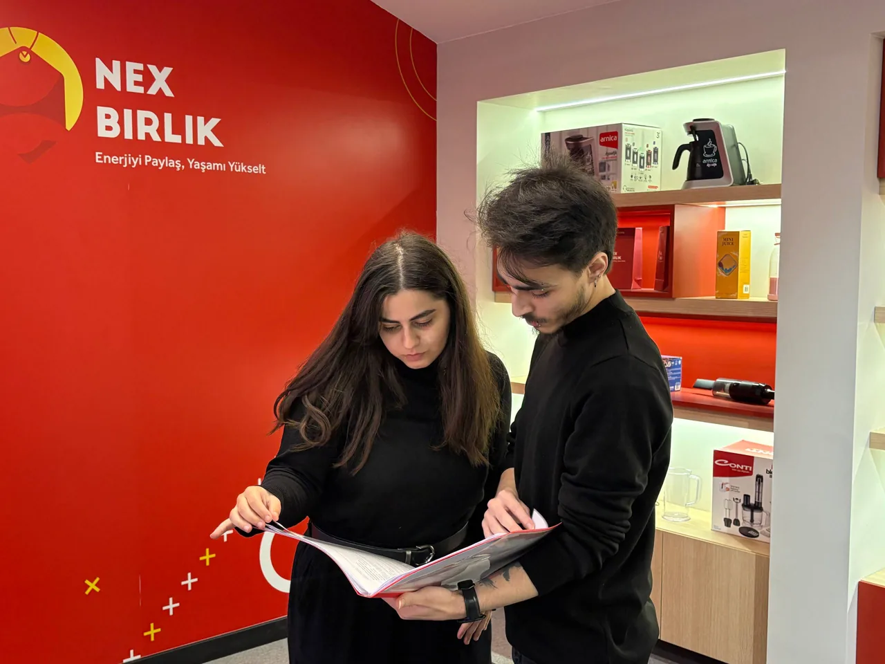 Kırmızı marka duvarının önünde materyale bakan iki kişi